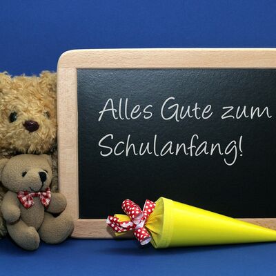 Zwei Teddys sitzen neben einer kleinen Schultafel, auf der geschrieben steht "alles Gute zum Schulanfang"