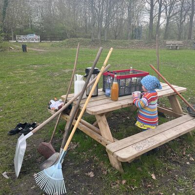 Pause beim Frühjahrsputz: Rechen, Schaufeln und andere Werkzeuge lehnen an einem Picknicktisch, ein Kind sitzt daneben mit einem Getränk, im Hintergrund eine grüne Wiese.