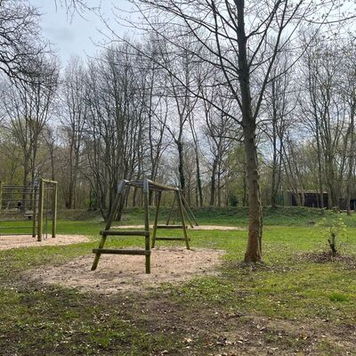 Bild vergr&ouml;&szlig;ern: Ein Spielplatz im Wald mit verschiedenen Klettergerüsten aus Holz auf Sandflächen. Die Bäume im Hintergrund haben noch wenig Laub, was auf den frühen Frühling hindeutet. Das Gelände ist von einer weiten Rasenfläche umgeben.
