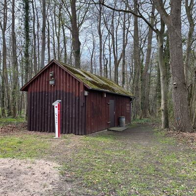 Bild vergr&ouml;&szlig;ern: Eine rustikale, dunkelbraune Holzhütte mit Satteldach am Rande eines Waldes. Direkt davor steht eine auffällige, weiß-rote Notrufsäule mit der Aufschrift „SOS“. Die Umgebung ist durch hohe Bäume und den sandigen Boden des angrenzenden Spielplatzes geprägt.
